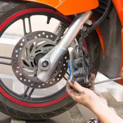 Mecánico ajustando el freno duro de una moto naranja. Se ve la rueda, el disco y la pinza de freno.