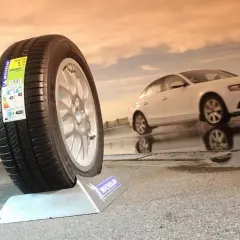 Neumático Michelin en primer plano, mostrando varios tipos de ruedas de coche. Un coche blanco se desplaza en el fondo sobre asfalto mojado.