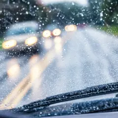 Gotas de lluvia en el parabrisas, luces borrosas de otros coches y la carretera mojada. Conducir con lluvia requiere precaución.