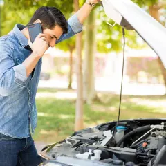 Hombre preocupado llama por teléfono junto a un coche con el capó abierto, revisando el **rendimiento del motor**.