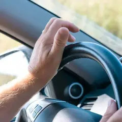 Mano sujetando el volante, con el brazo peludo visible. La dirección del coche dura se siente firme mientras el conductor avanza.