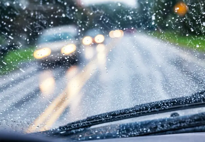 Gotas de lluvia en el parabrisas, luces borrosas de otros coches y la carretera mojada. Conducir con lluvia requiere precaución.