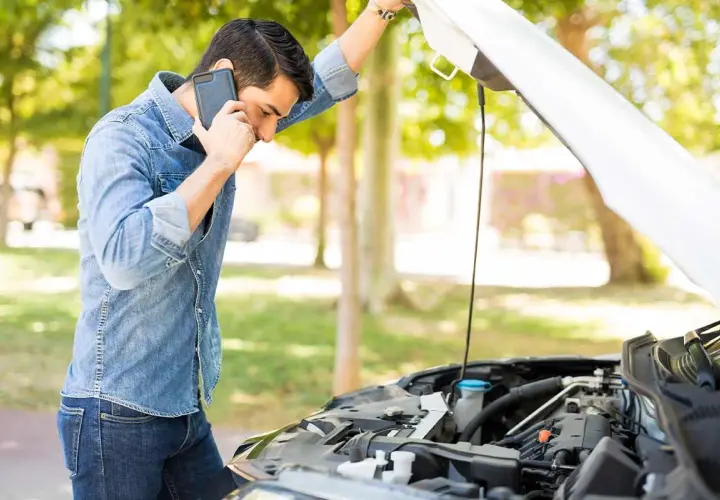 Hombre preocupado llama por teléfono junto a un coche con el capó abierto, revisando el **rendimiento del motor**.