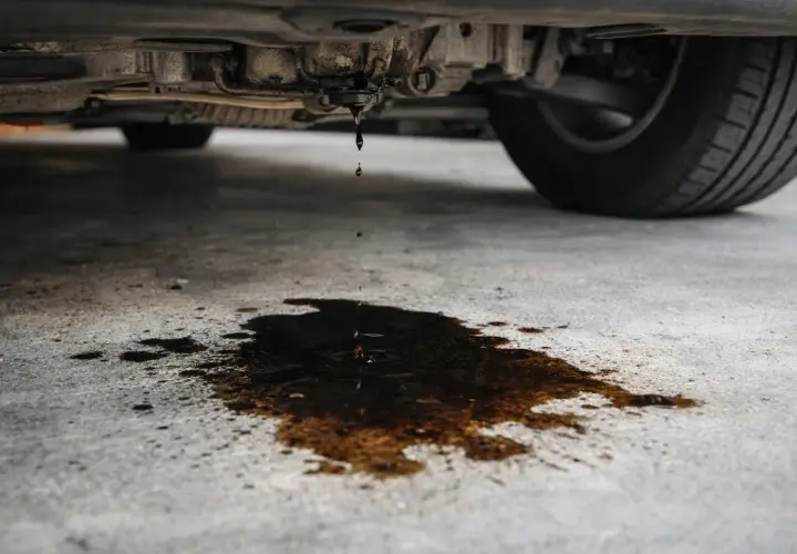 Got it. Here's the alt text in Spanish:

Aceite goteando del coche. Mi coche pierde aceite por debajo, dejando un charco oscuro en el suelo de cemento.