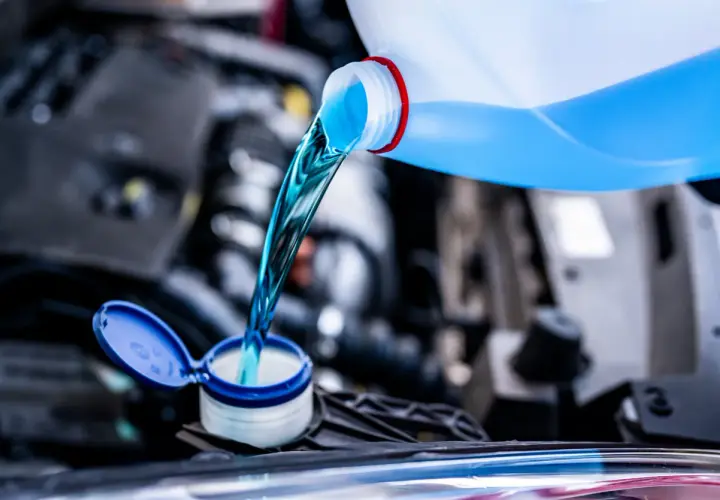 Líquido azul brillante se vierte de una botella a un depósito del coche. Es hora de decidir qué anticongelante usar para proteger tu motor.