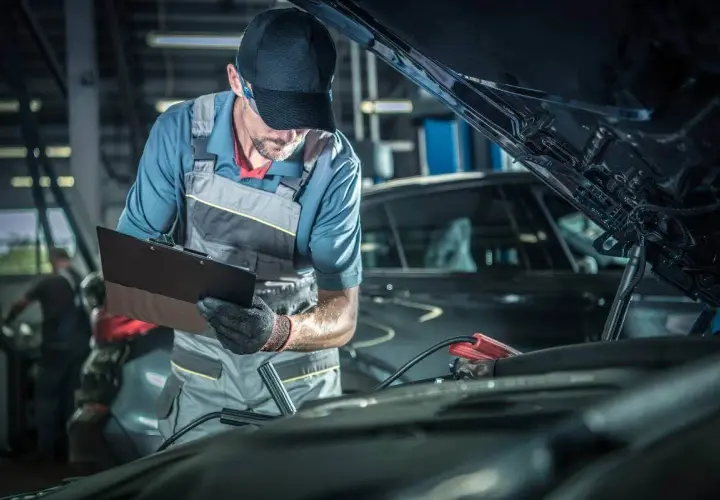 Mecánico con gorra y mono realiza la revisión del coche, anotando detalles en una carpeta. Cables de arranque conectados al motor.