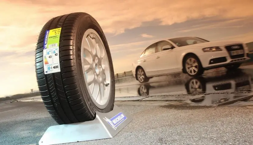 Neumático Michelin en primer plano, mostrando varios tipos de ruedas de coche. Un coche blanco se desplaza en el fondo sobre asfalto mojado.