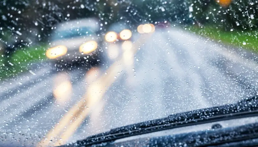 Gotas de lluvia en el parabrisas, luces borrosas de otros coches y la carretera mojada. Conducir con lluvia requiere precaución.