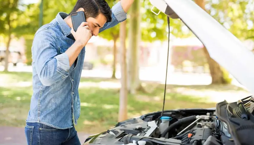 Hombre preocupado llama por teléfono junto a un coche con el capó abierto, revisando el **rendimiento del motor**.