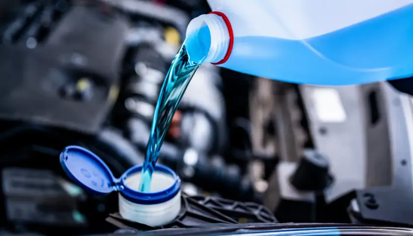 Líquido azul brillante se vierte de una botella a un depósito del coche. Es hora de decidir qué anticongelante usar para proteger tu motor.