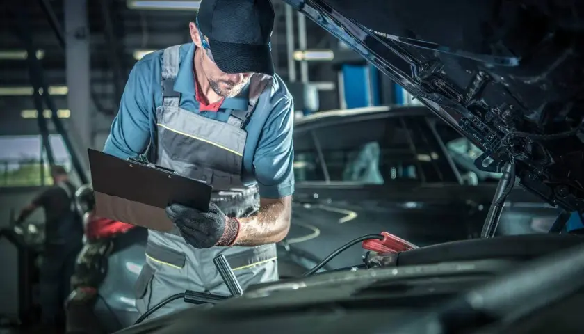 Mecánico con gorra y mono realiza la revisión del coche, anotando detalles en una carpeta. Cables de arranque conectados al motor.