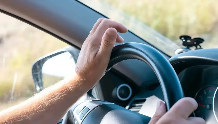 Mano sujetando el volante, con el brazo peludo visible. La dirección del coche dura se siente firme mientras el conductor avanza.