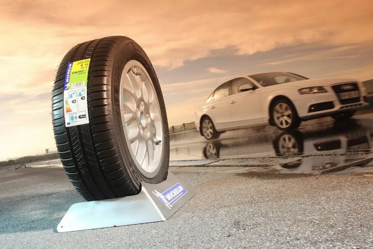 Neumático Michelin en primer plano, mostrando varios tipos de ruedas de coche. Un coche blanco se desplaza en el fondo sobre asfalto mojado.