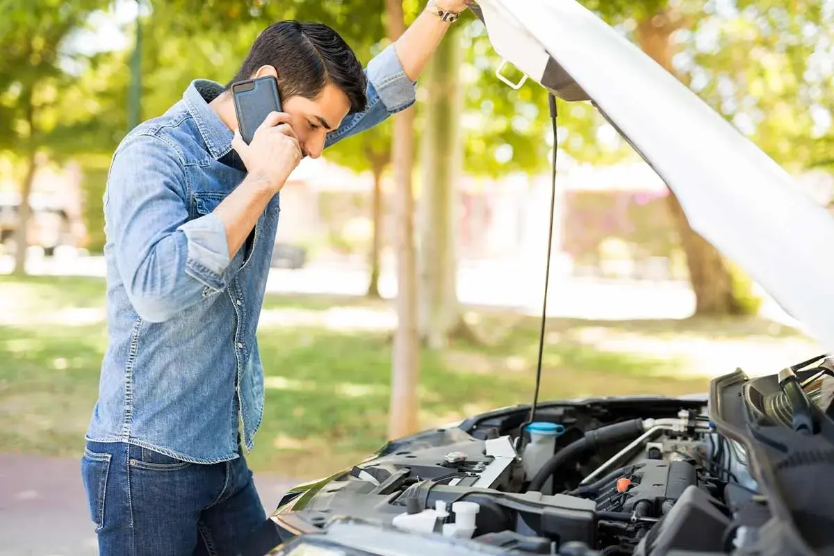 Hombre preocupado llama por teléfono junto a un coche con el capó abierto, revisando el **rendimiento del motor**.