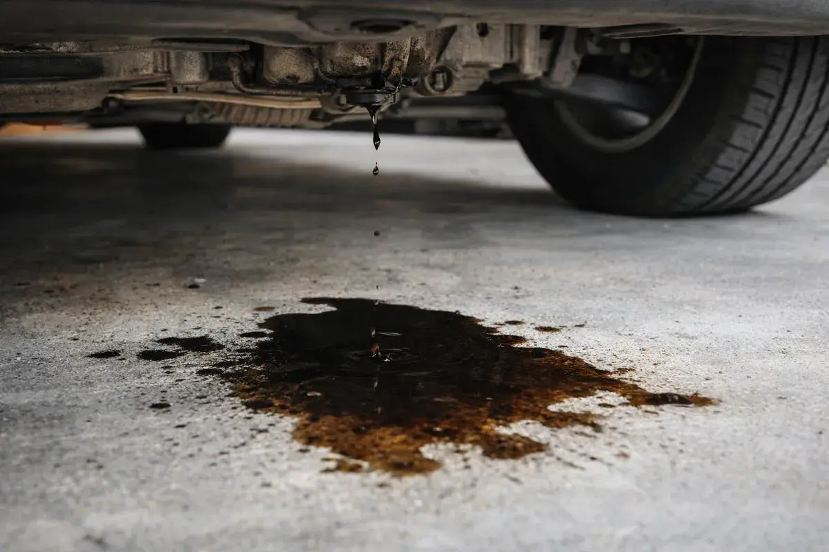Got it. Here's the alt text in Spanish:

Aceite goteando del coche. Mi coche pierde aceite por debajo, dejando un charco oscuro en el suelo de cemento.
