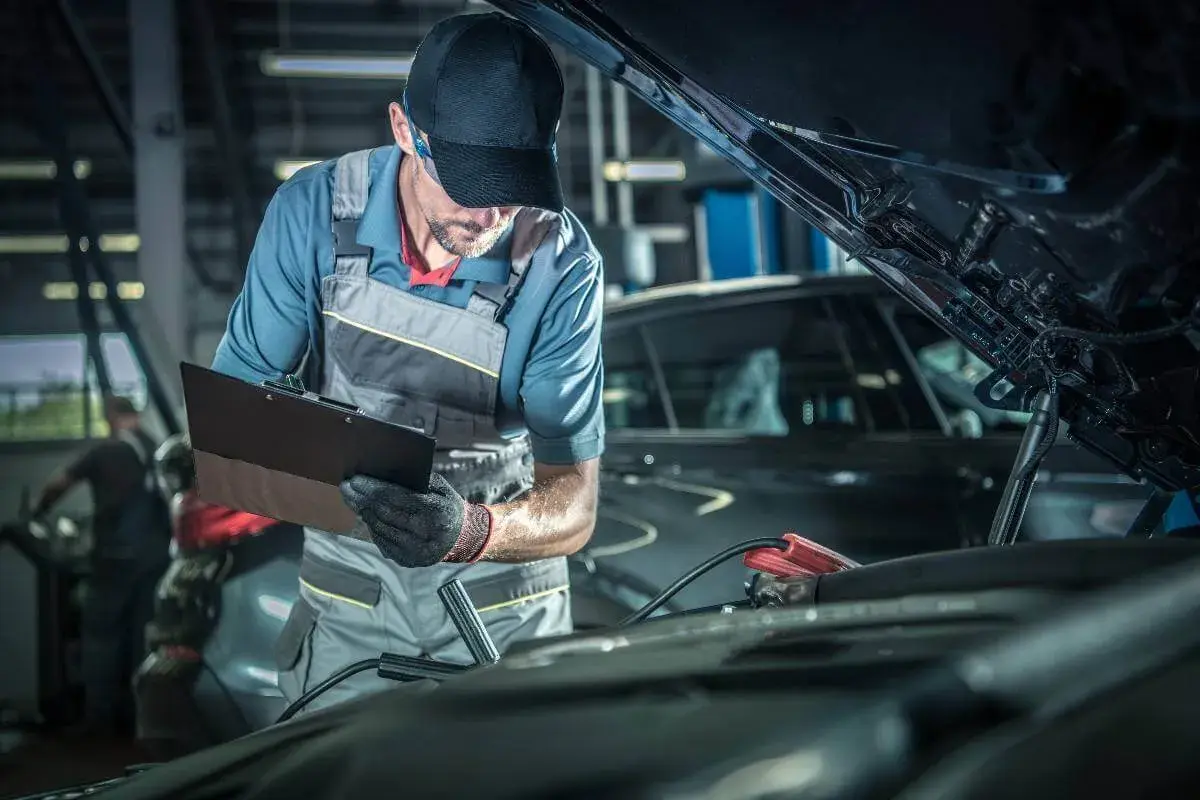 Mecánico con gorra y mono realiza la revisión del coche, anotando detalles en una carpeta. Cables de arranque conectados al motor.