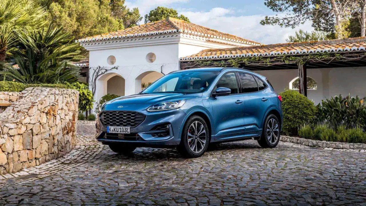 Ford Kuga PHEV, un SUV azul aparcado en un camino empedrado frente a una casa blanca. A pesar de su apariencia, algunos usuarios reportan ford kuga phev problemas.