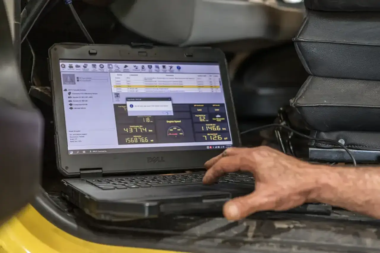 Mano revisando datos en laptop Dell para optimizar el rendimiento del motor. Muestra horas de motor, odómetro y consumo de combustible.
