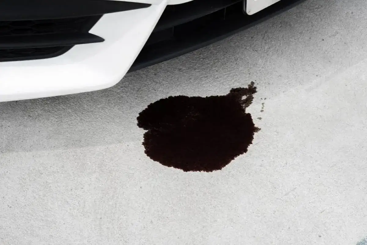 Mancha oscura de aceite en el suelo, justo debajo de la parte delantera de un coche blanco. Parece que mi coche pierde aceite por debajo.